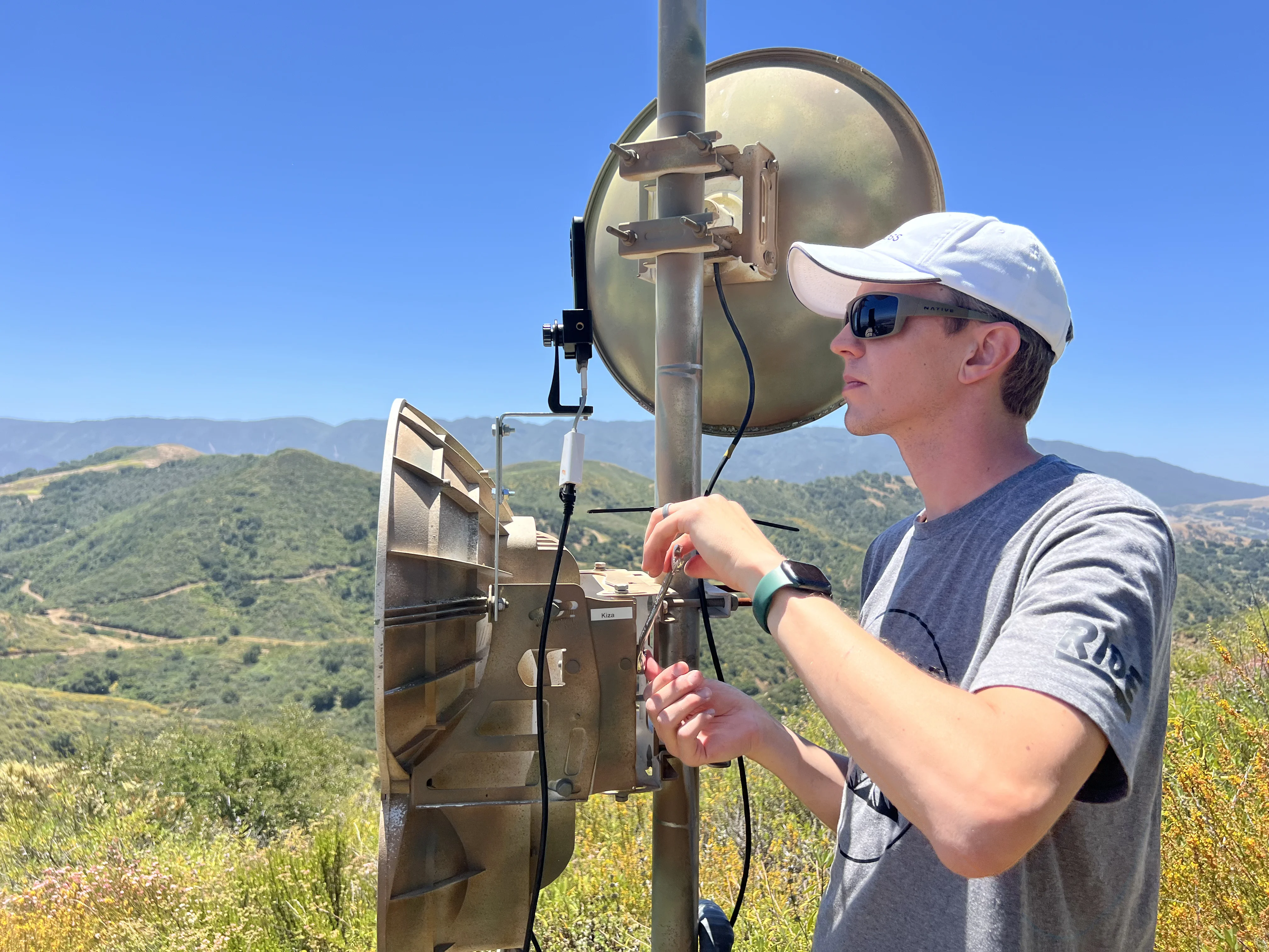 Technician adjusting AirFiber 24 with phone mounted, hilltop installation with mountain valley behind